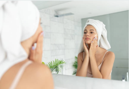 A woman wraps her head with a towel, looks at herself in the mirror and gently touches her face to check how skincare products are being absorbed after treatment with a microneedle patch.