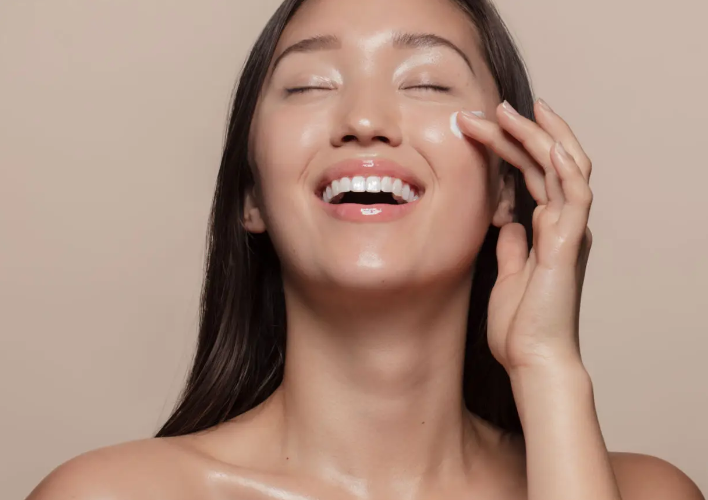 A woman with long, dark hair smiles brightly as she applies moisturizer to her face.